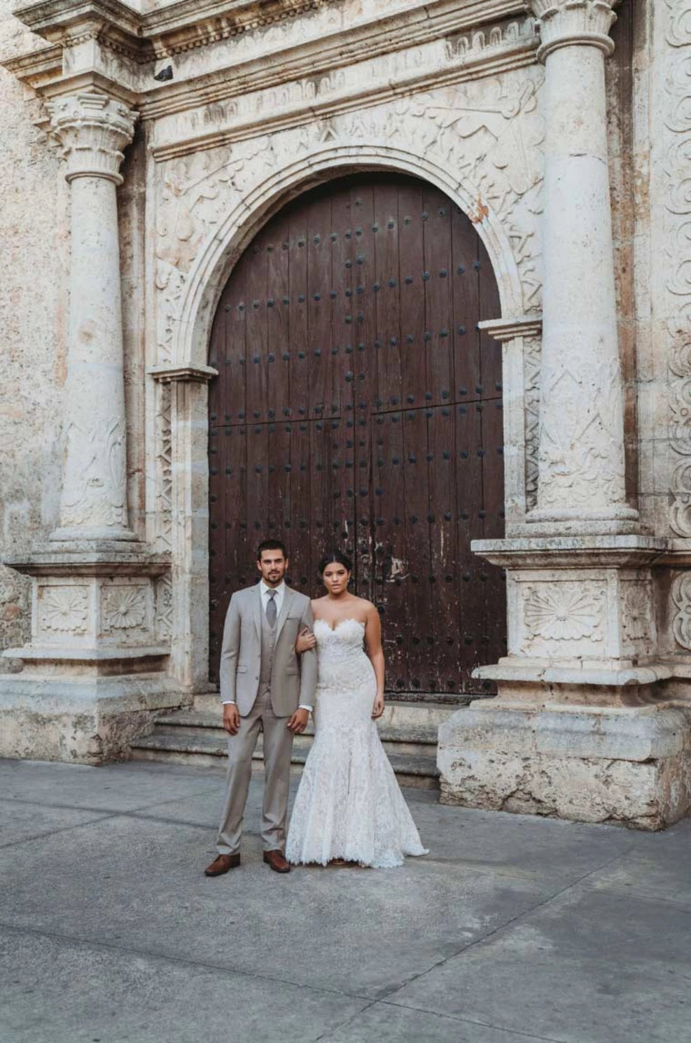 Groom in sand suit with a bride standing in front of a large stone archway in Mexico.