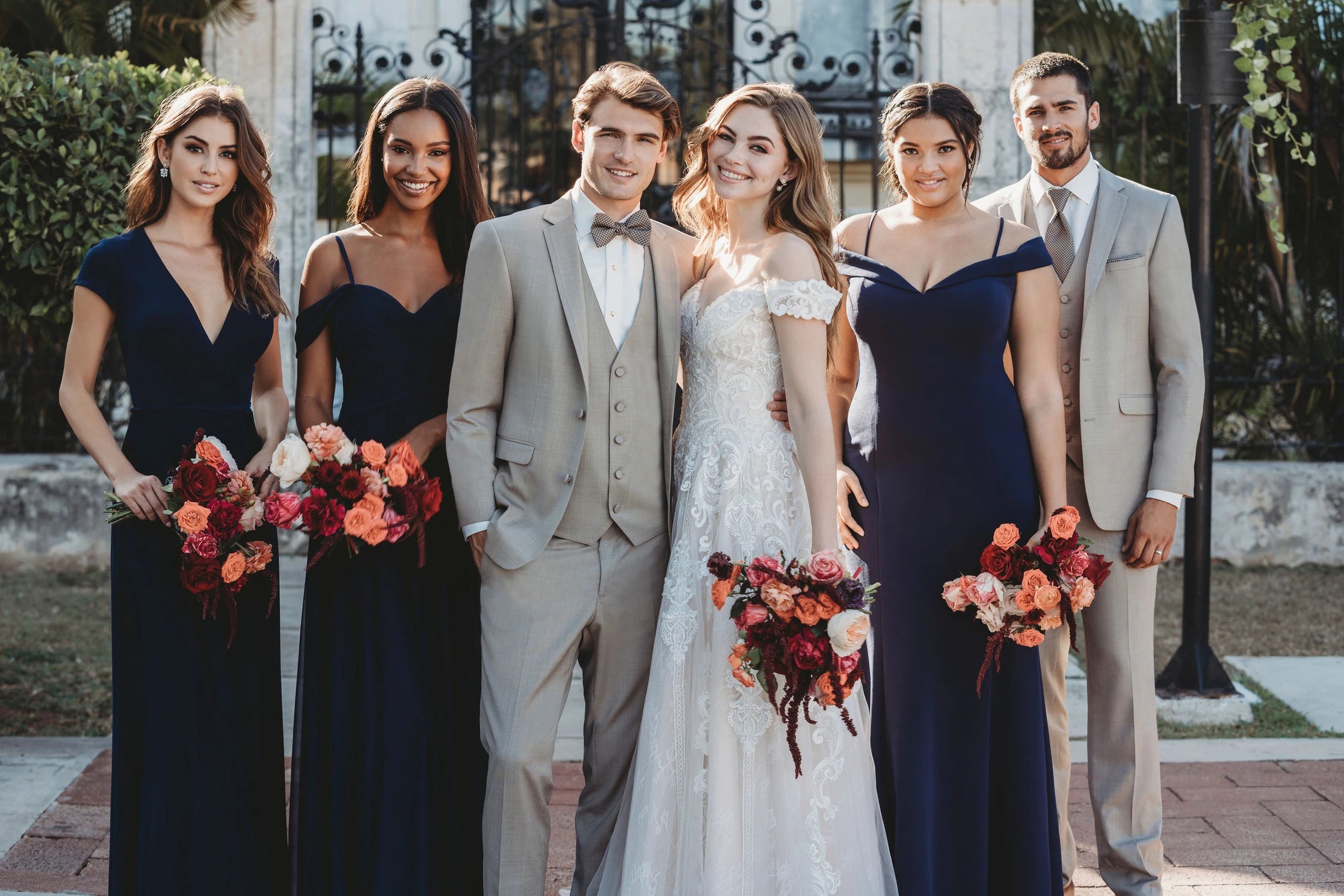 groom wearing a sand suit and bowtie accompanied by a best man and 3 bridesmaids wearing navy blue