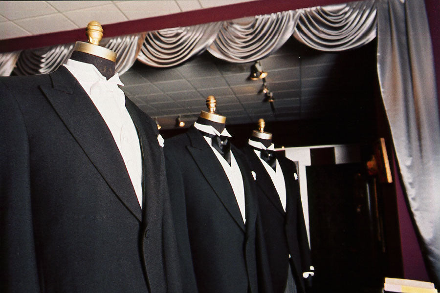 Row of black cutaway morning suits on mannequins in a tuxedo shop with curtains in the background.