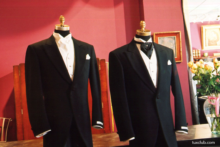 Two black morning suits on mannequins against a red wall with decorative elements.
