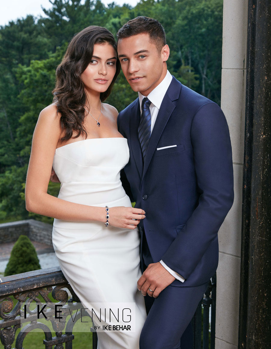 Man in a navy suit and woman in a white dress standing together outdoors | Tuxclub