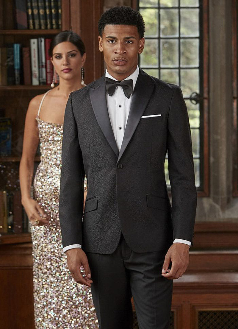 Man in a black brocade tuxedo standing next to a woman in a sparkling dress indoors.