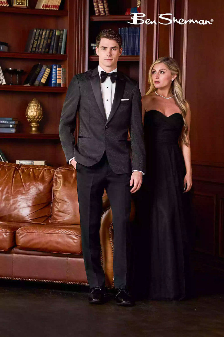 Man in black paisley tuxedo and woman in black evening gown standing in a room with bookshelves and a brown leather couch.