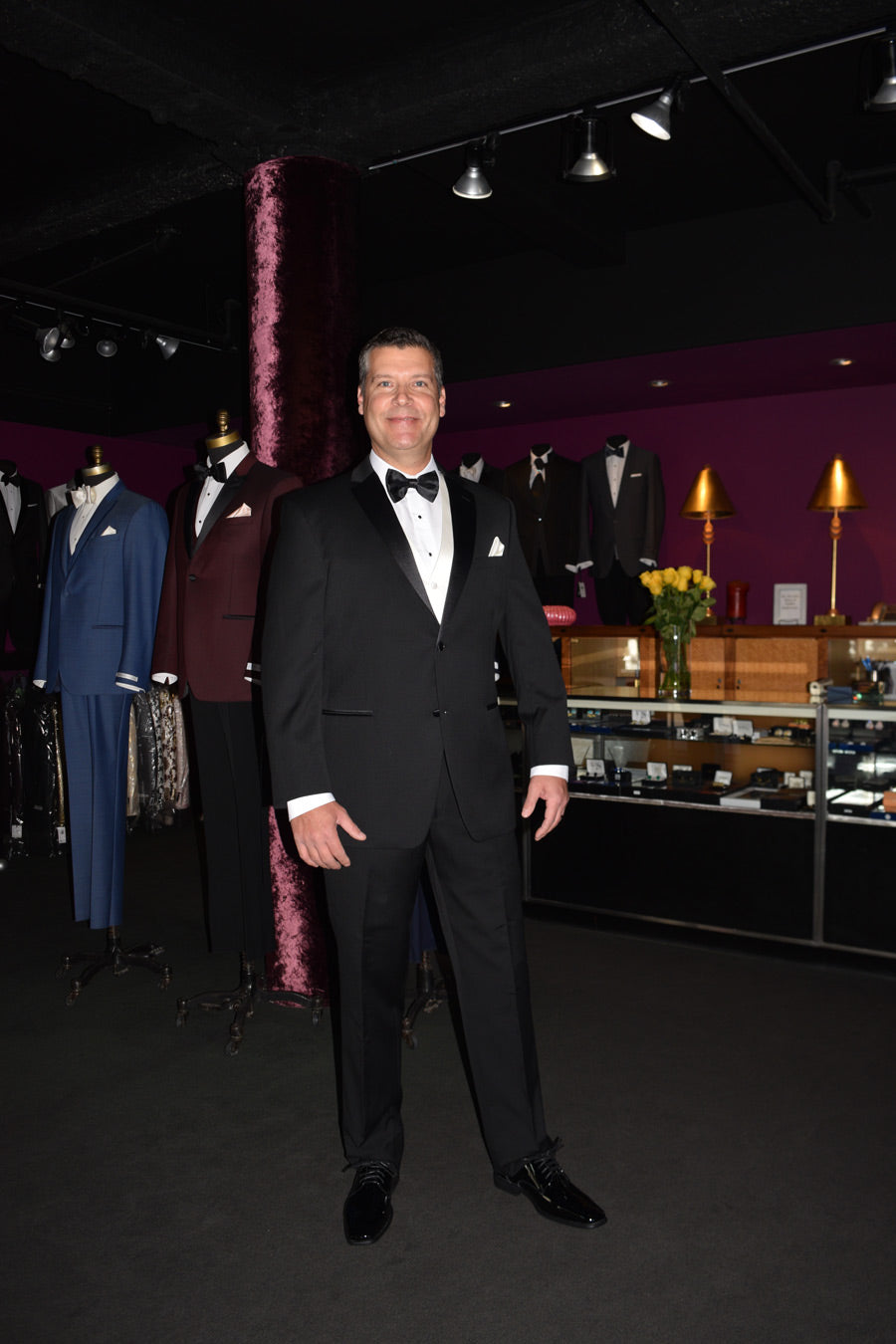 Man wearing a black slim-fit notch lapel tuxedo with white vest and black satin bowtie at a tuxedo shop
