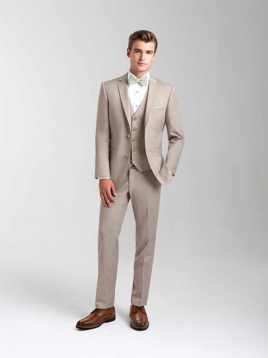 30 year old caucasian model wearing a sand suit and bowtie with brown shoes standing in front of a gray background