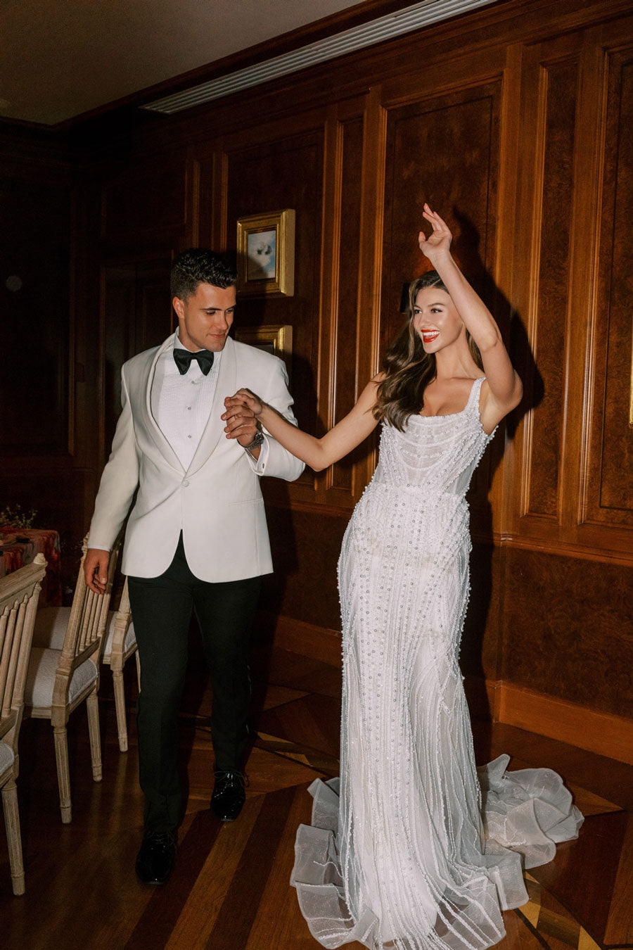 Groom in an off-white shawl collar tuxedo and a bride in a off-white dress dancing in a dining room with wooden paneling.