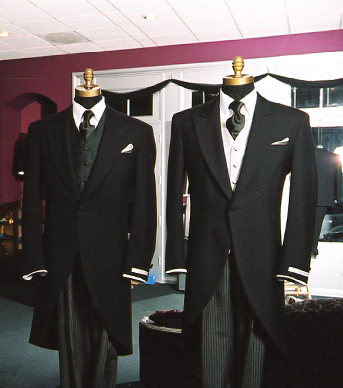 Two black morning suits on mannequins against a pink wall.