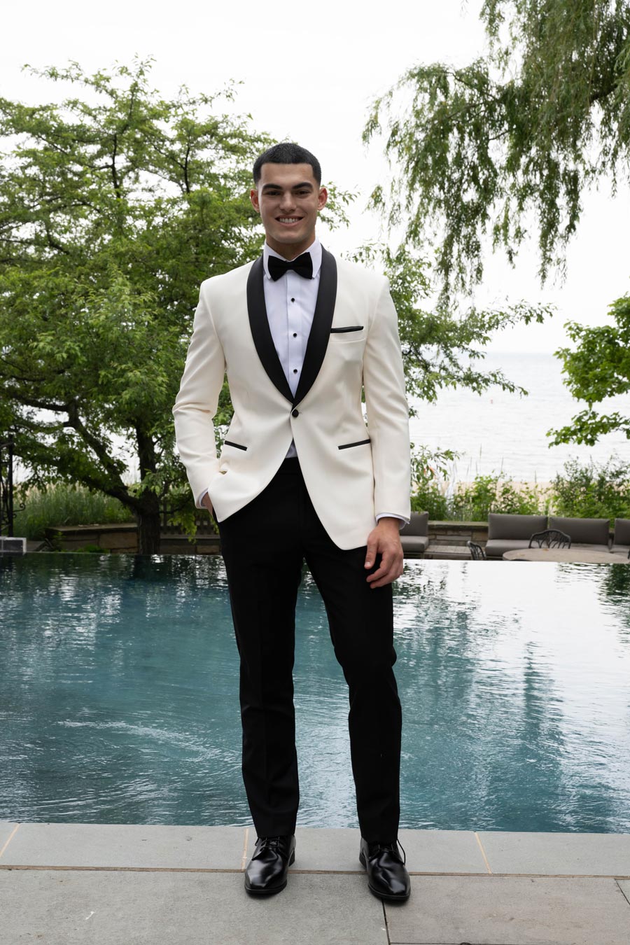 Younger hispanic man wearing an ivory tuxedo with black shawl collar standing by a pool with trees.