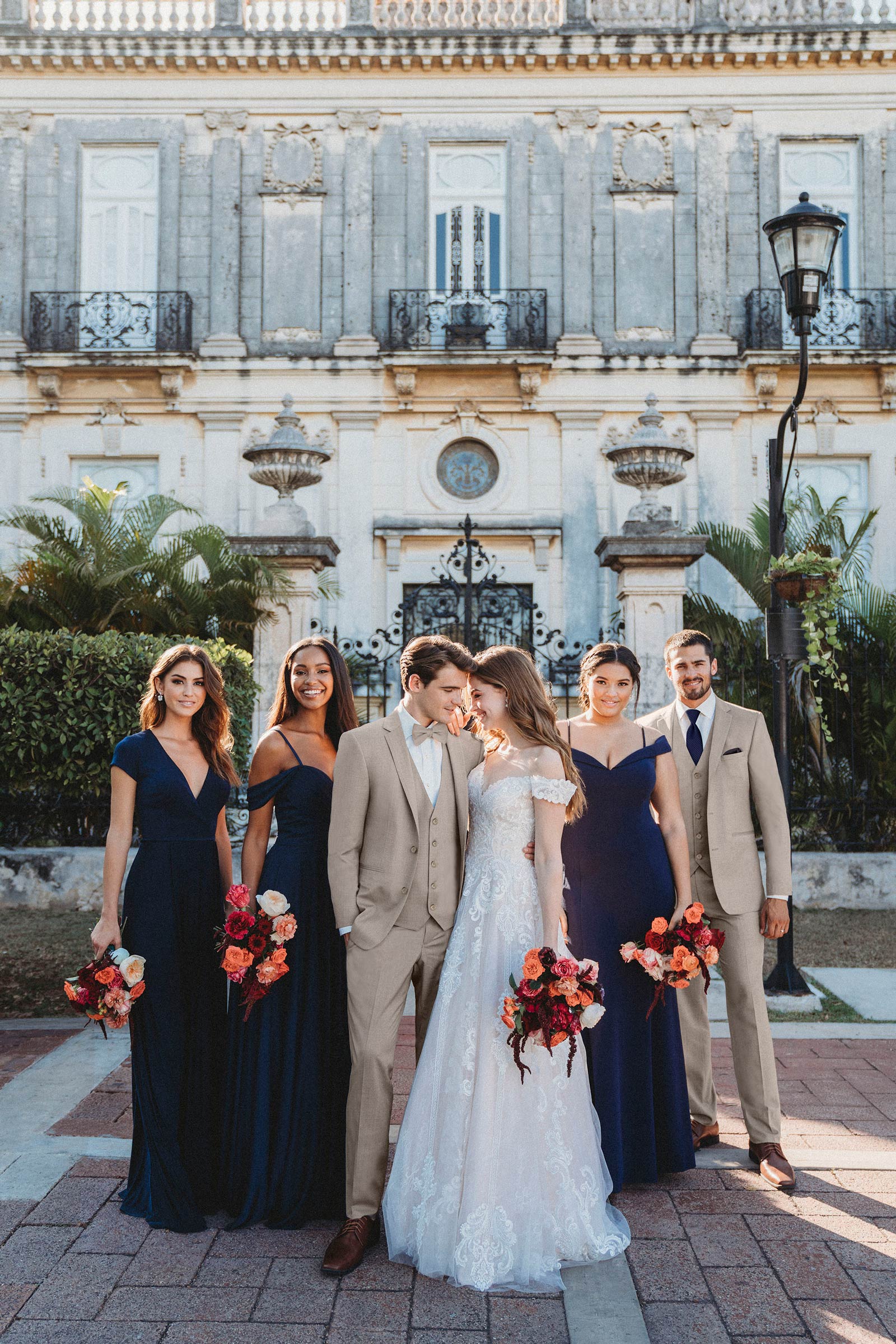group photo of a wedding party where the men are wearing the sand suit, with bridesmaids wearing long navy blue gowns.