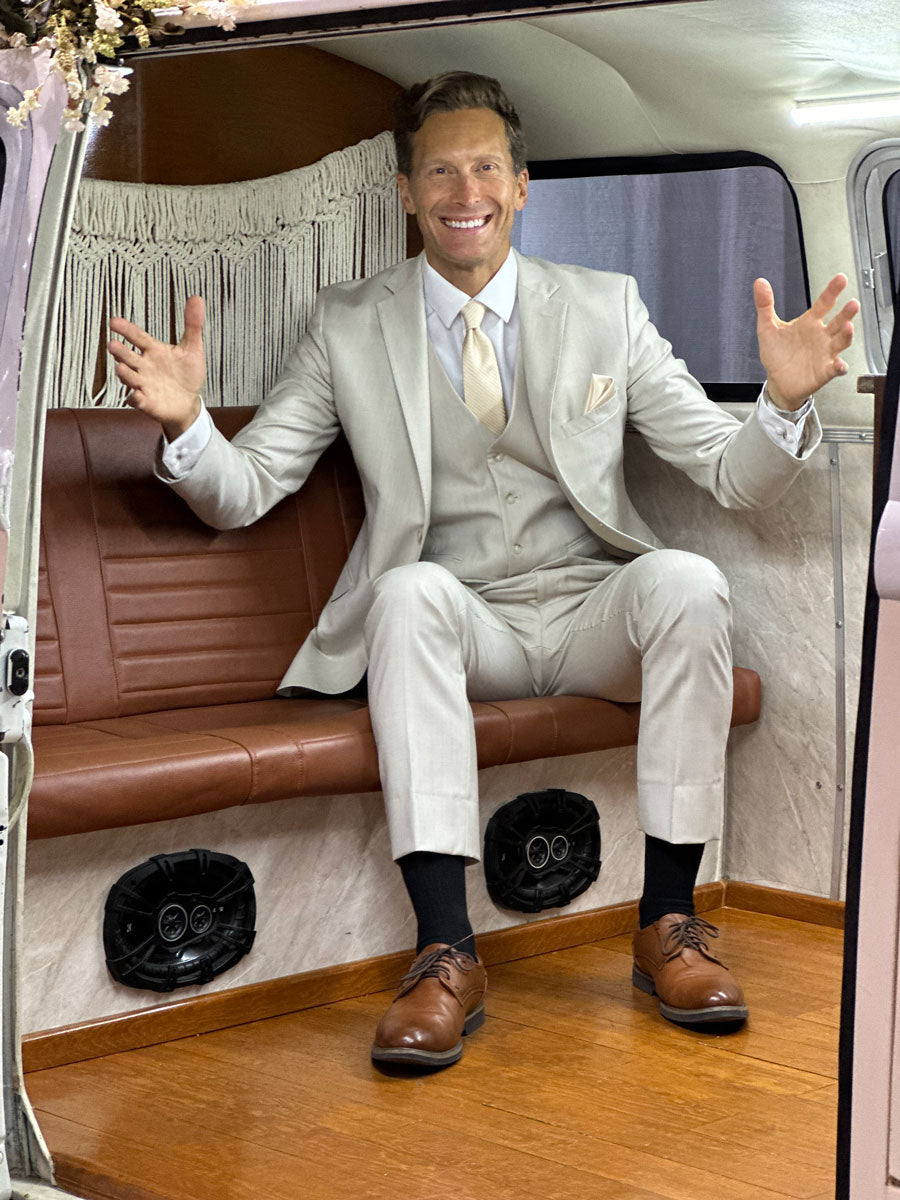 Man wearing a tan fitted suit while sitting in the back of a vintage van at a wedding reception.