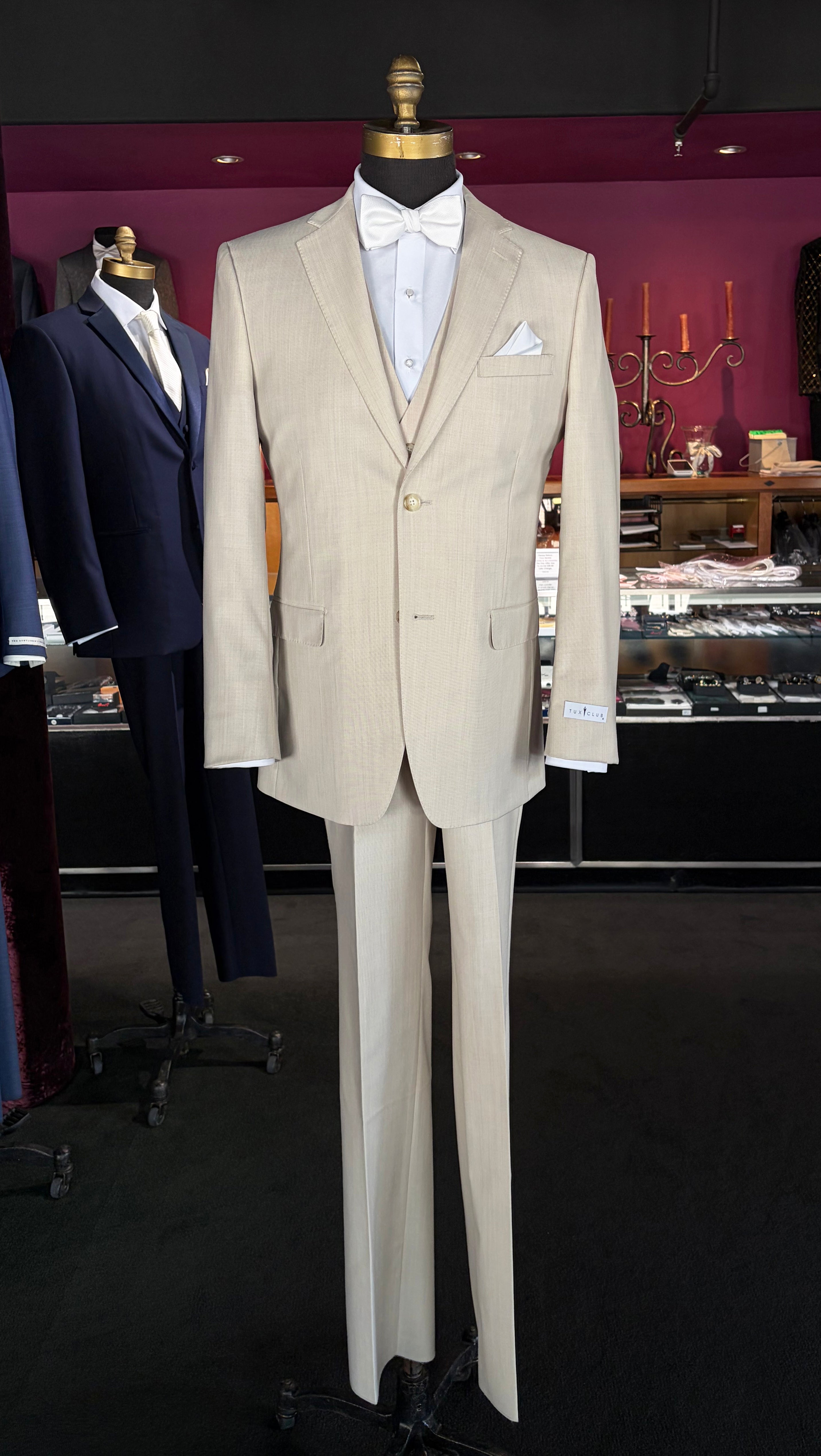 A tan fitted two-piece suit displayed on a mannequin torso, styled with a white dress shirt, white bow tie, and white pocket square, set against a dark boutique interior.