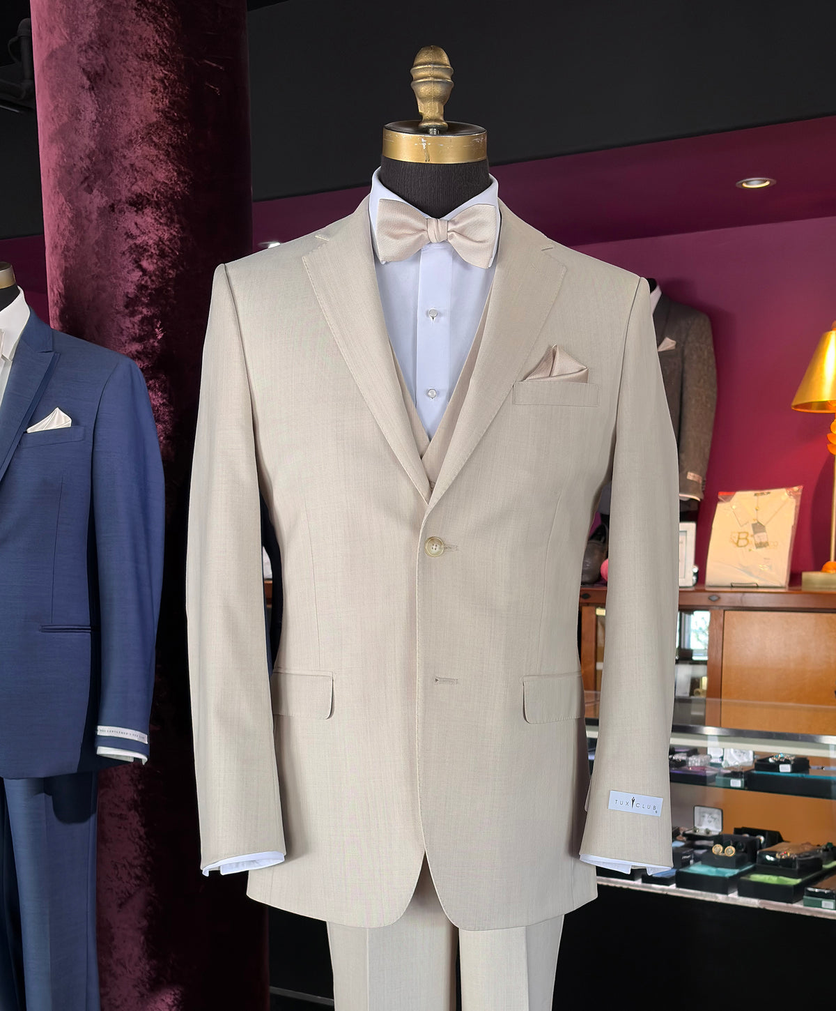 close-up tan fitted three-piece suit displayed on a mannequin, featuring a jacket, matching trousers, a white shirt, a beige bow tie, and a pocket square, set against a dark background inside a tuxedo shop