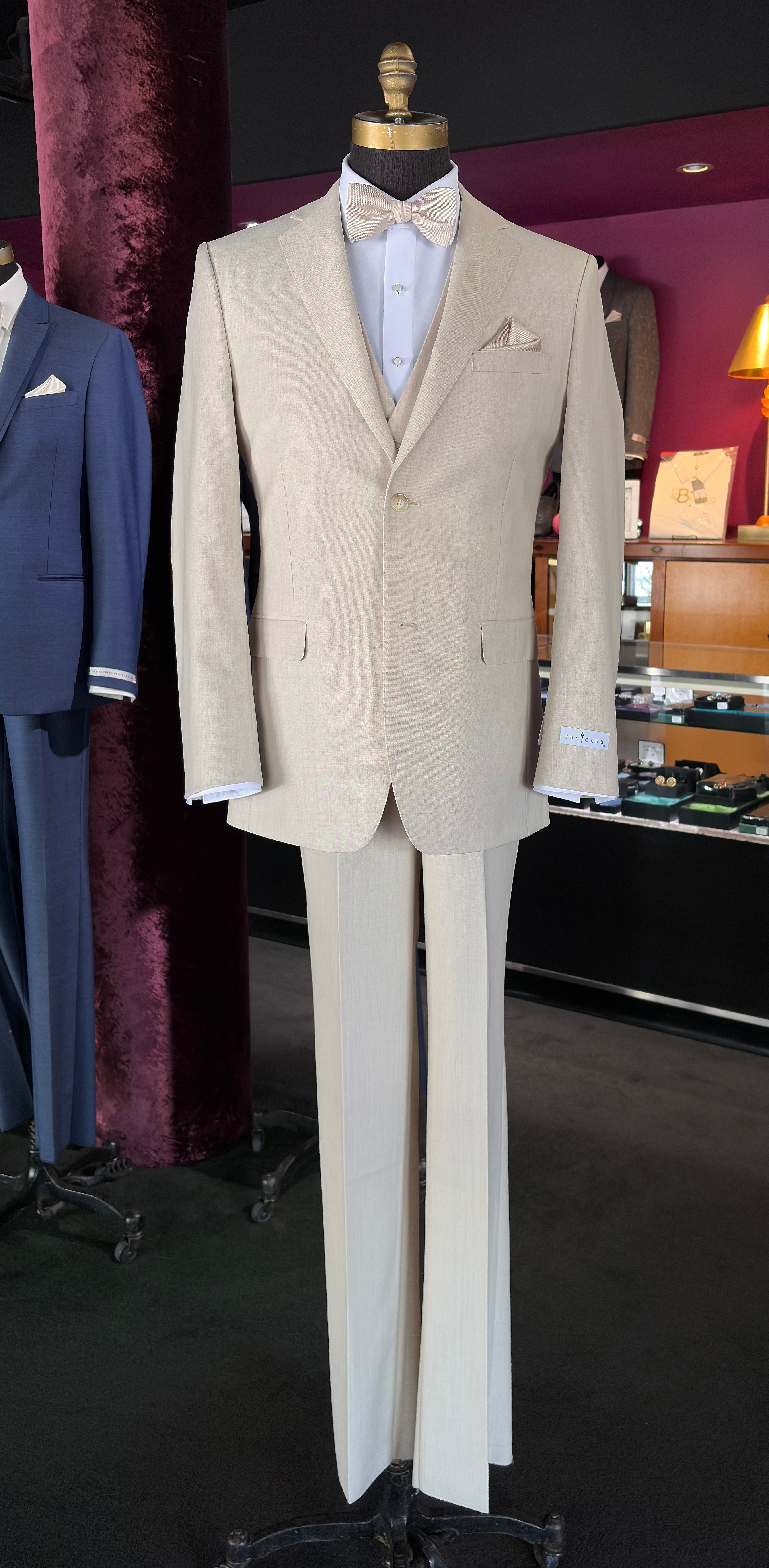 A tan fitted three-piece suit displayed on a mannequin, featuring a jacket, matching trousers, a white shirt, a beige bow tie, and a pocket square, set against a dark background inside a tuxedo shop