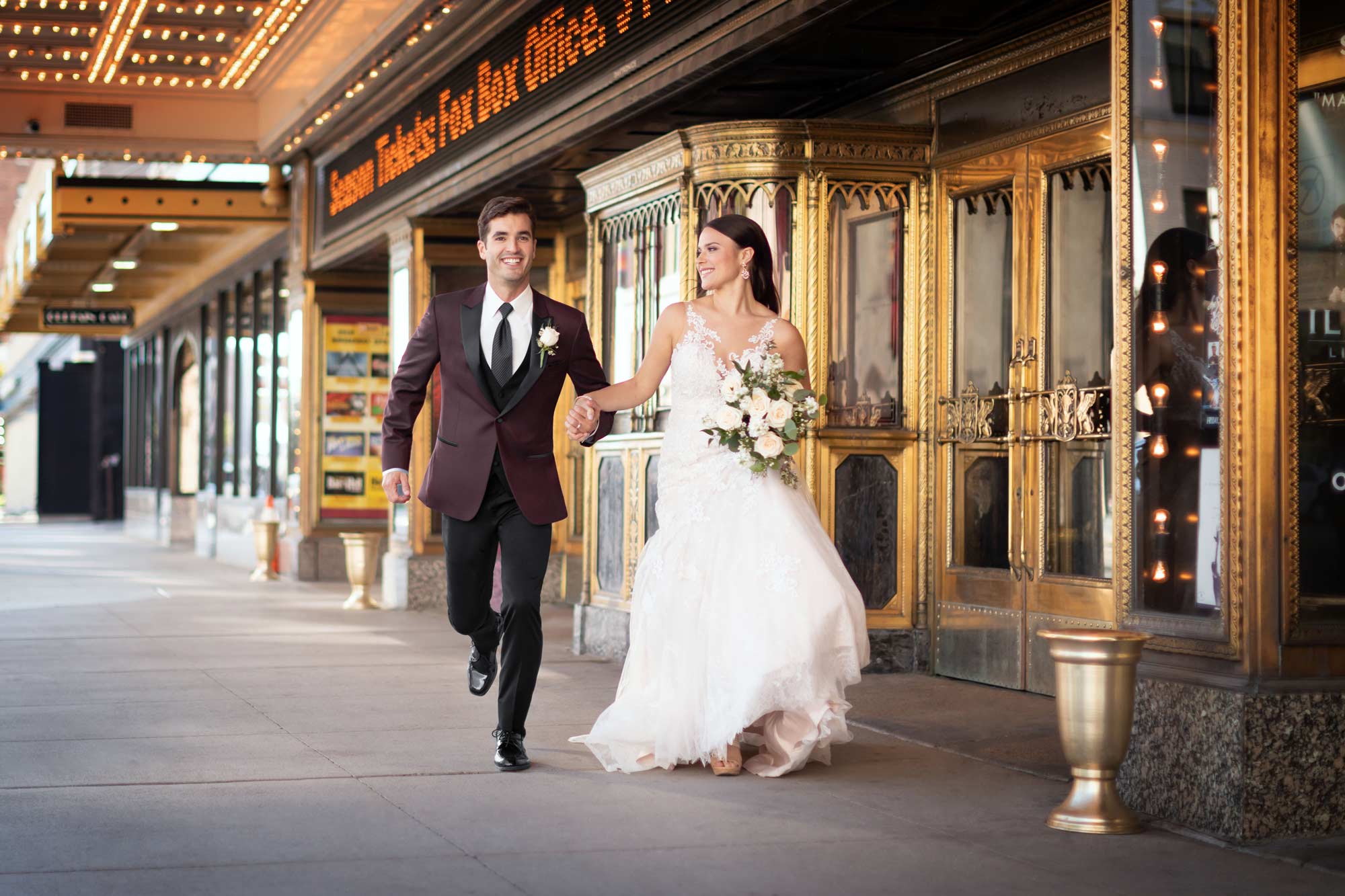 Man in burgundy tuxedo and woman in a wedding gown walking in front of a theater entrance.