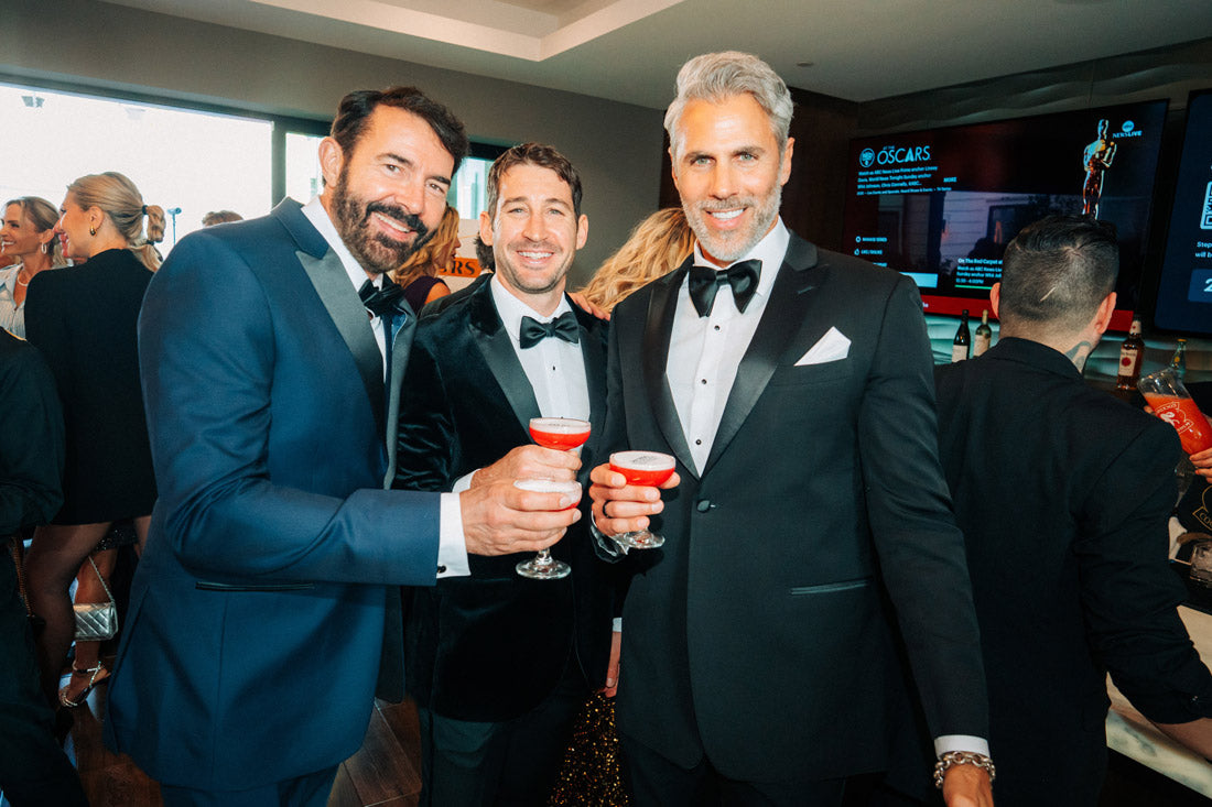 A man wearing a black peak lapel tuxedo at an Oscars awards celebration in La Jolla, California standing with 2other men in tuxedos.