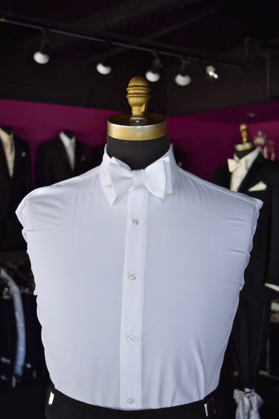 Mannequin wearing a white tuxedo shirt with a white pique "self-tie" bowtie inside a tuxedo shop with other tuxedos in the background