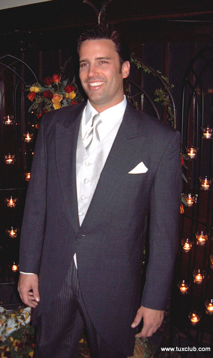 man wearing a charcoal cutaway morning suit with silver vest and silver long dress tie against a wall with candles