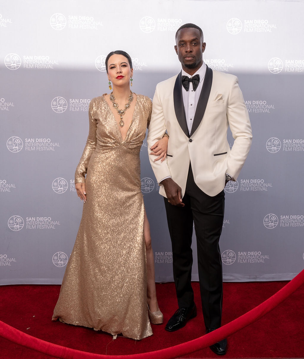 A black man wearing an ivory tuxedo with black lapel from tuxclub with a woman in a champagne gown on a red carpet at the San Diego International Film Festival.