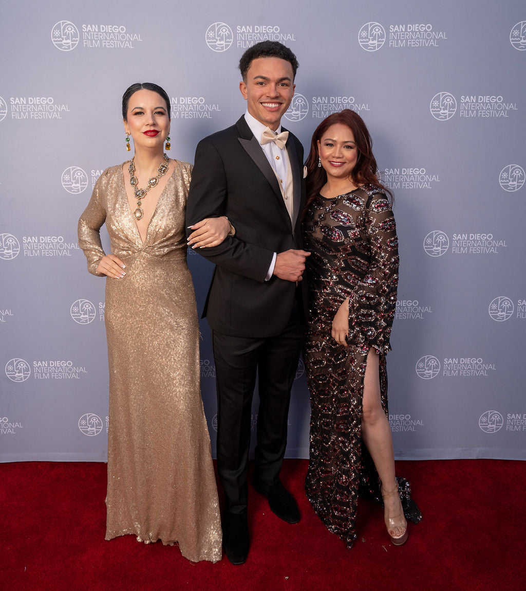 man is wearing a black peak lapel tuxedo with a champagne colored bowtie and vest with two women standing on red carpet
