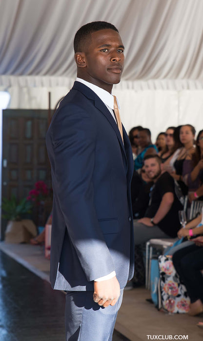 side view close up black man in a navy blue slim suit walking on a runway with an audience in the background.