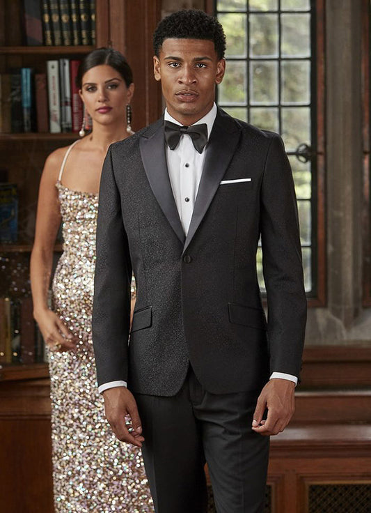 Man in a black brocade tuxedo standing next to a woman in a sparkling dress indoors.