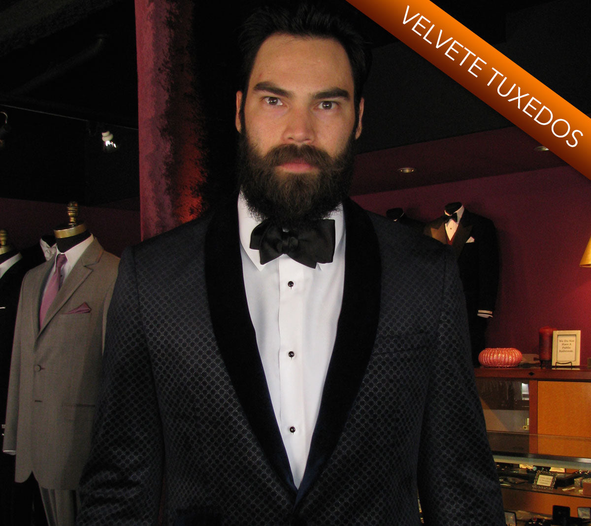 Man wearing a black velvet shawl collar tuxedo with a bow tie in a clothing store setting.
