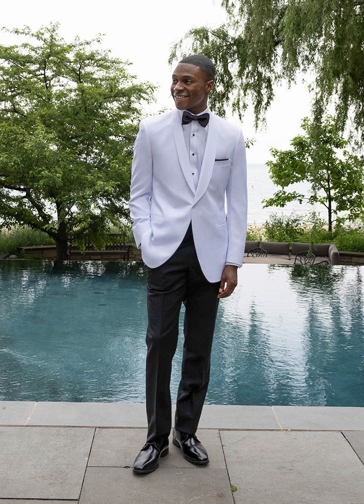 Man in a white shawl collar dinner jacket standing by a pool with greenery in the background