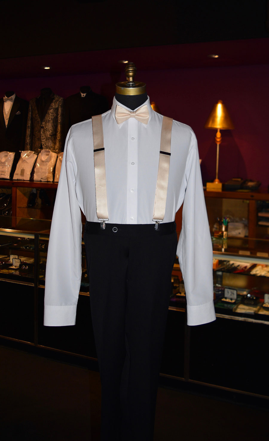 Mannequin wearing a white dress shirt with champagne satin suspenders and black pants in a tuxedo shop setting.