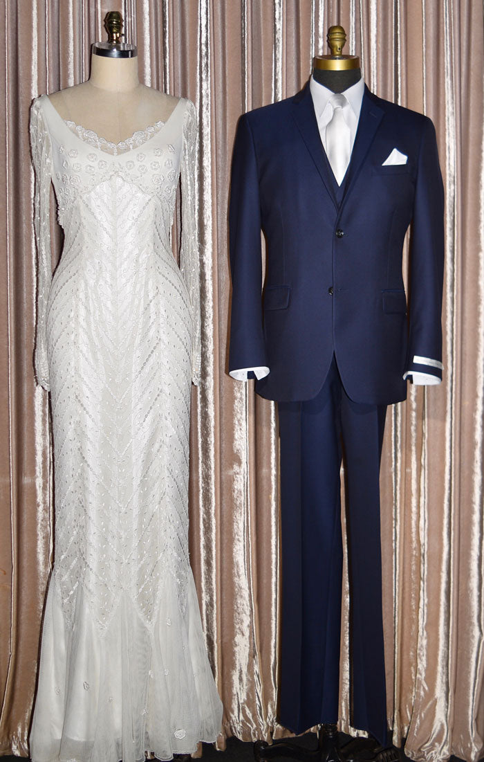Full length mannequin wearing a navy blue slim-fit suit on mannequins with a white dress tie next to a white lace dress