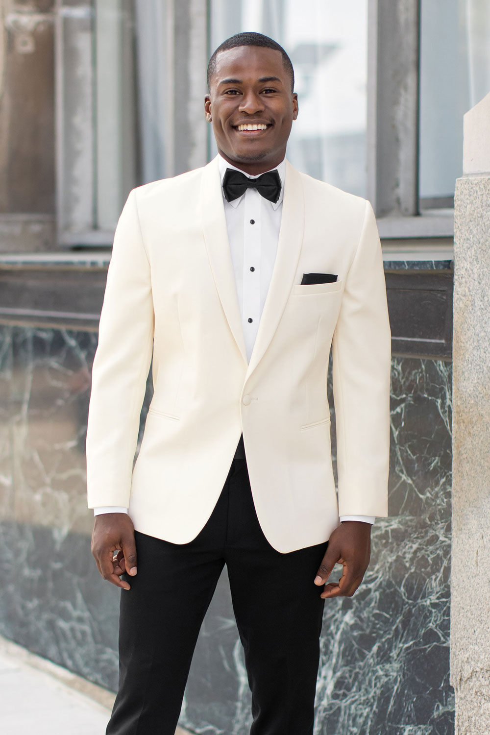 Handsome black man wearing an ivory shawl collar tuxedo with a black bow tie standing against a marble wall.