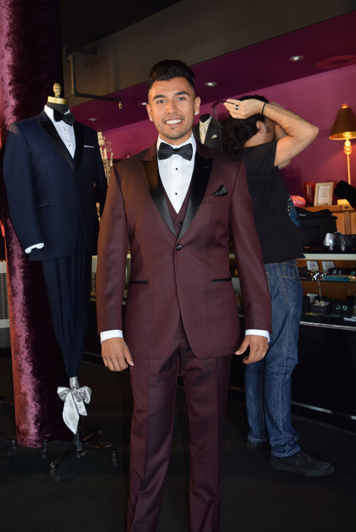 Man wearing a burgundy tuxedo with a white shirt and black bow tie, standing in a  tuxedo shop with suits on display.