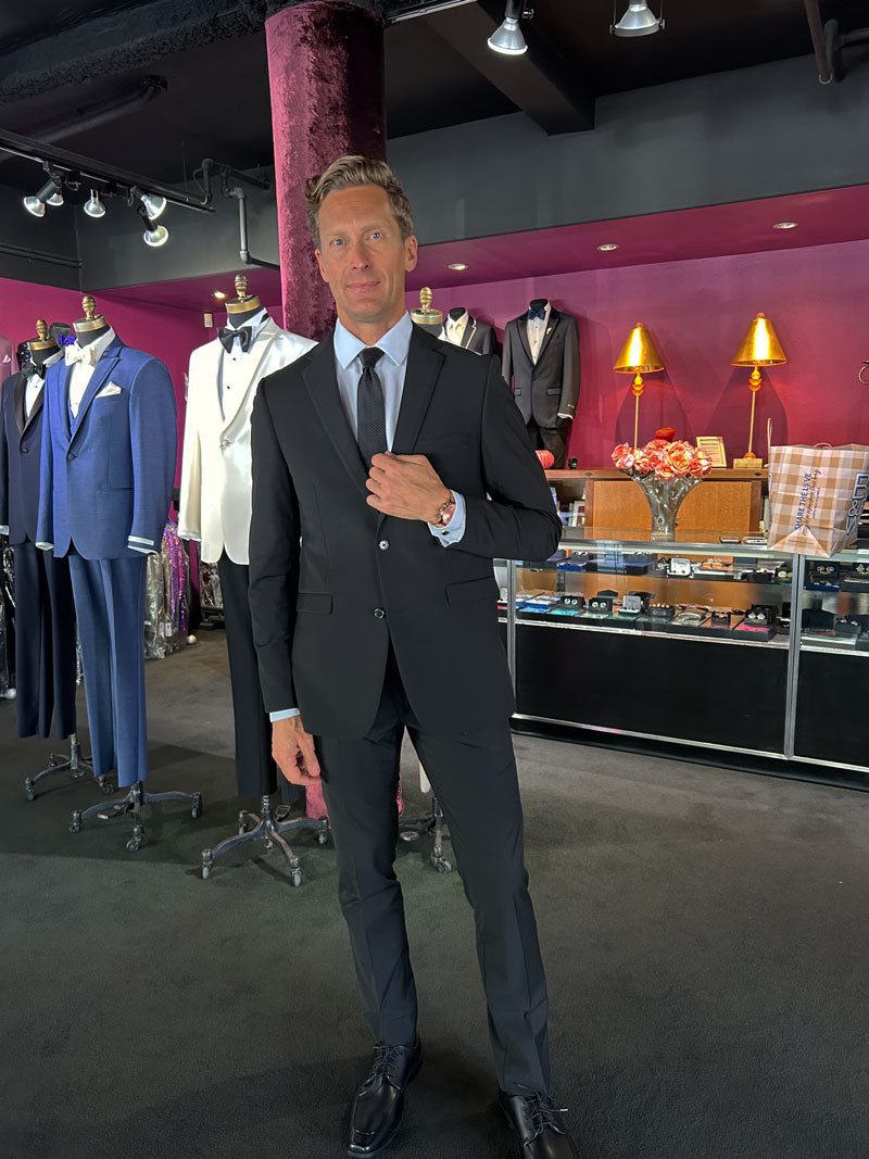 Man trying-on a black slim fit suit at a tuxedo shop with suits on display.