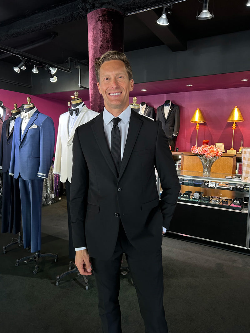 Man in a black slim-fit suit standing in a clothing store with suits and accessories on display.
