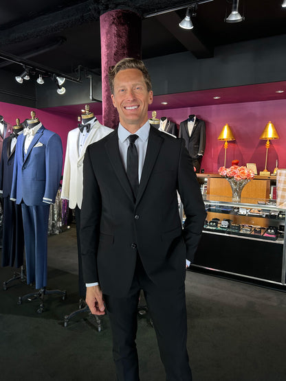 Man in a black slim-fit suit standing in a clothing store with suits and accessories on display.