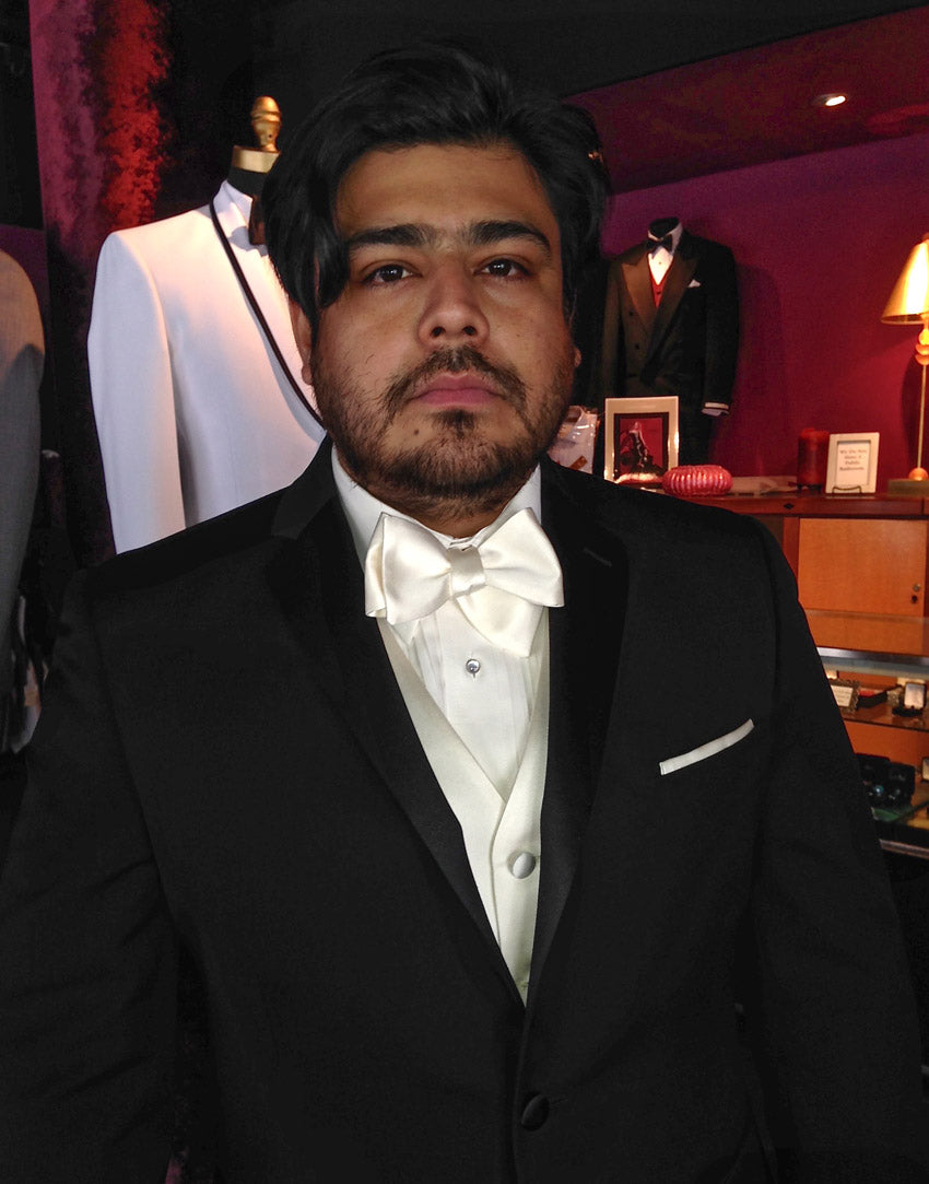 Man in a black tuxedo with a ivory bow tie and ivory satin tuxedo vest in a room with formal attire on display.