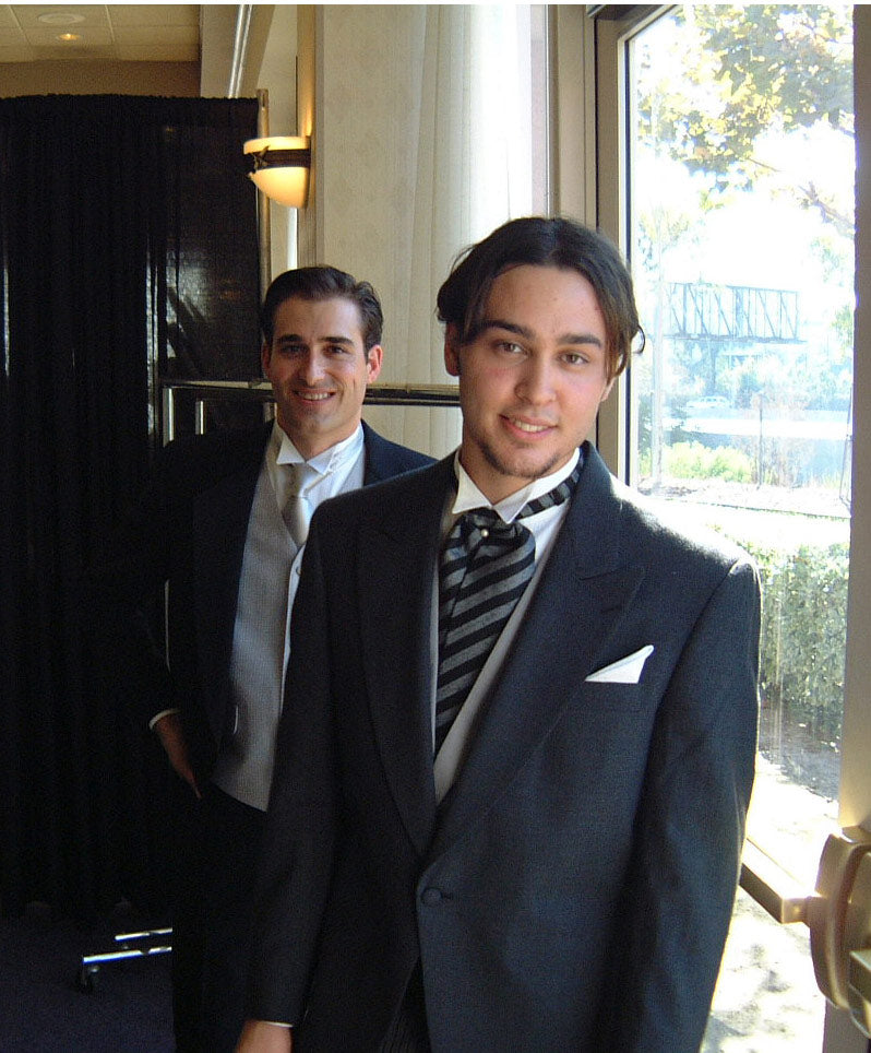 Two men in formal attire standing indoors , one wearing a charcoal cutaway morning suit style, with a big window in the background.