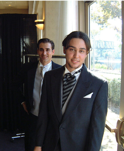 Two men in formal attire standing indoors , one wearing a charcoal cutaway morning suit style, with a big window in the background.