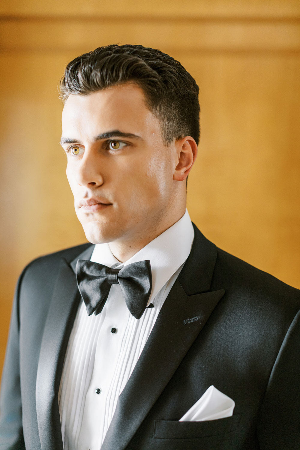 Close up of groom wearing a one button peak lapel tuxedo from TUXCLUB with black satin bowtie and a white tuxedo shirt with pleats. 