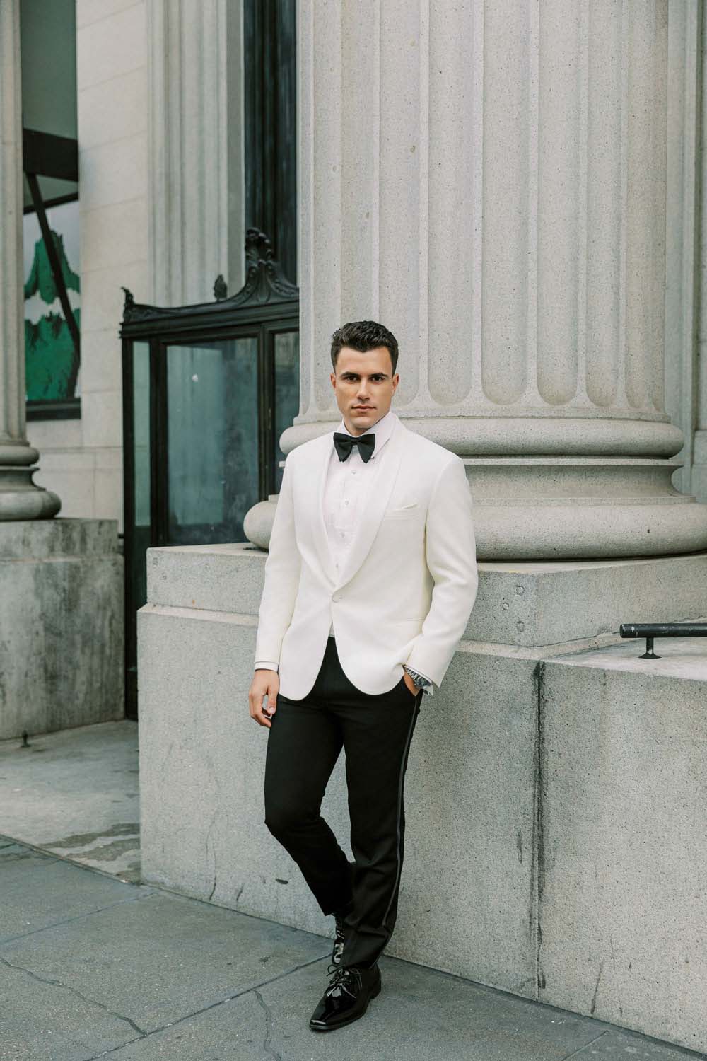 Groom wearing an ivory shawl collar tuxedo with black satin bowtie standing in front of a roman column historic building.