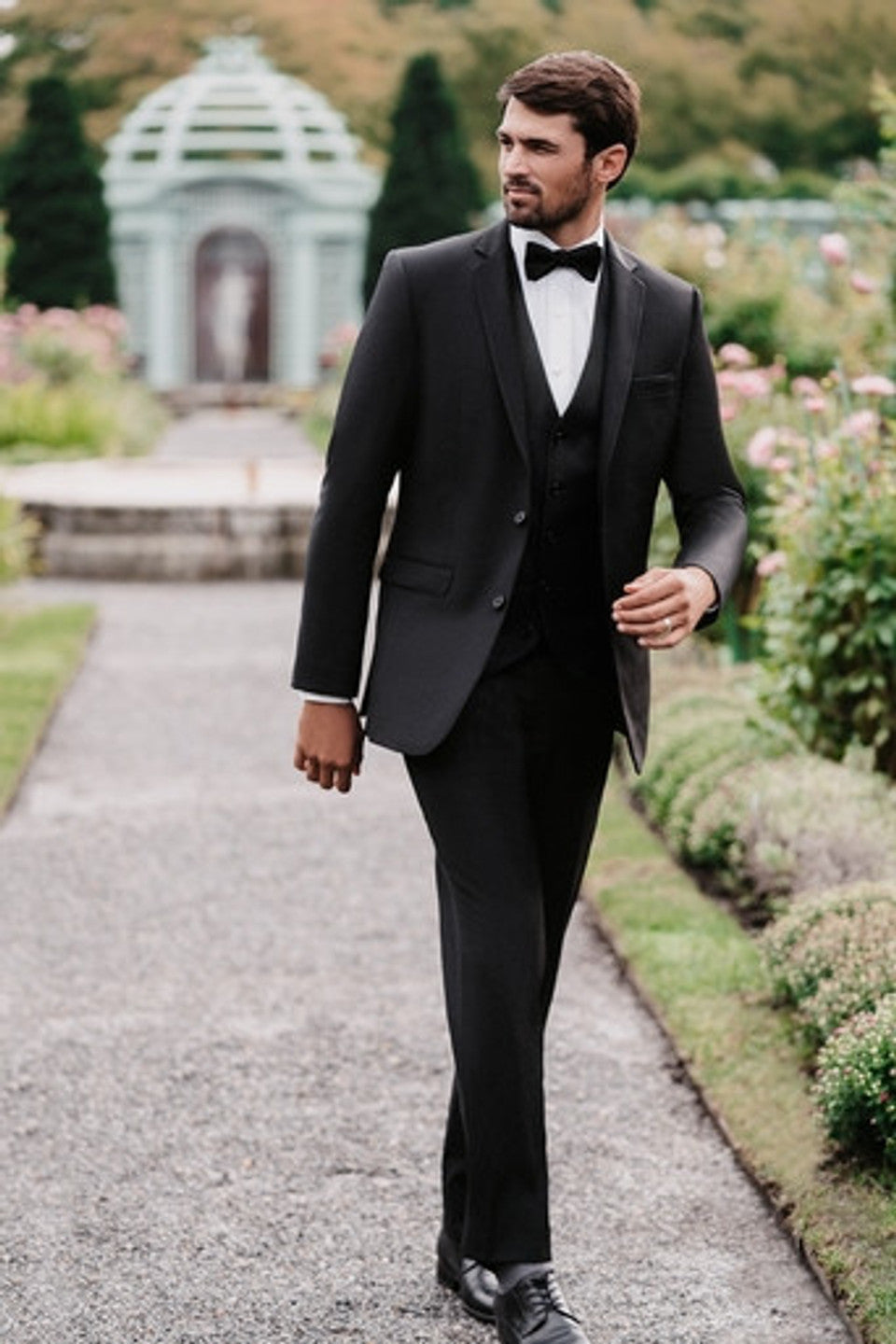 Man in a black slim-fit suit with black bowtie walking through a garden at a wedding