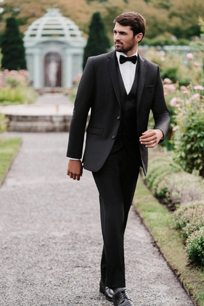 Man in a black slim-fit suit with black bowtie walking through a garden at a wedding