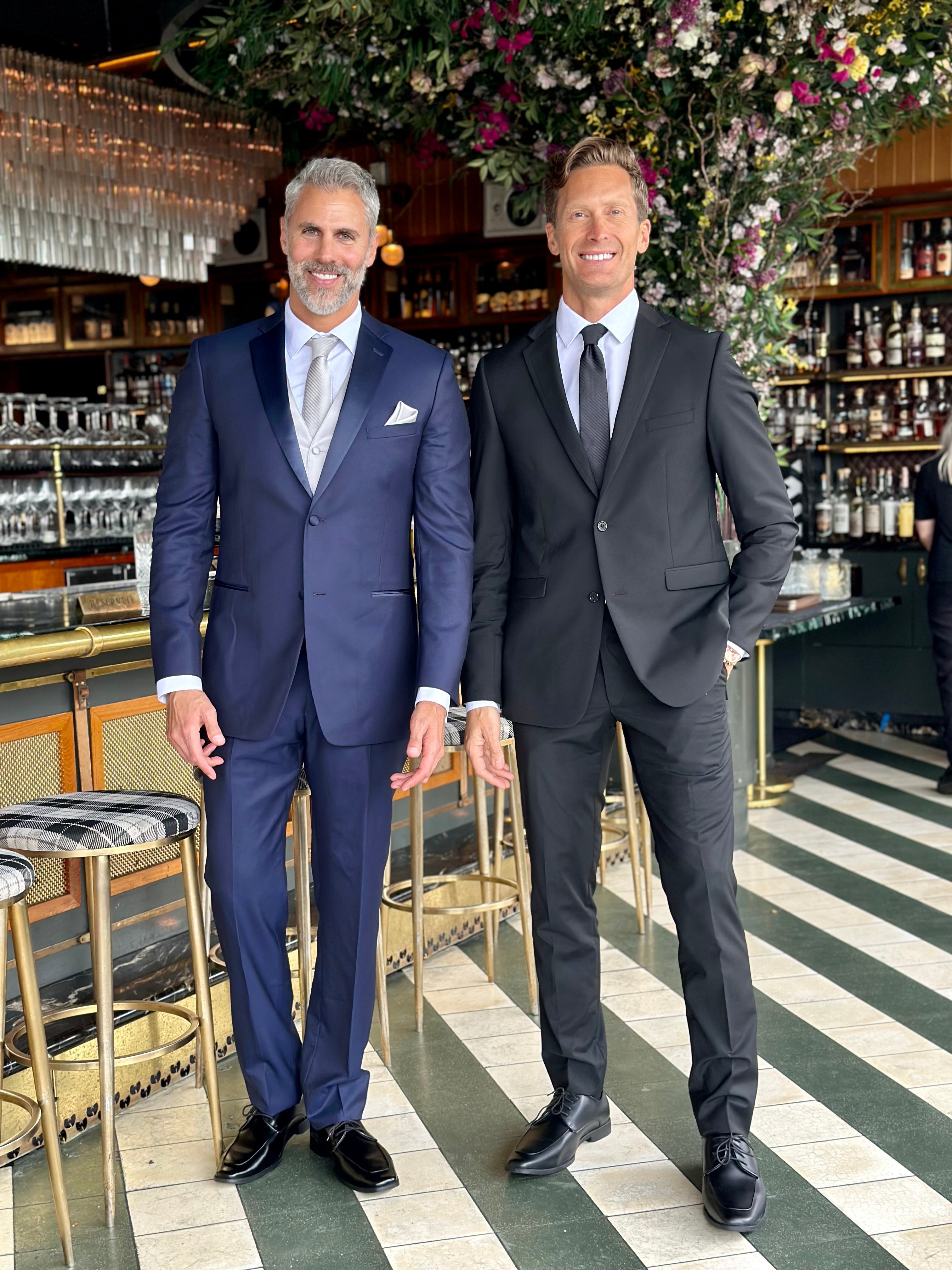 Man standing in a bar wearing a black slim-fit suit accompanied by a man wearing a blue slim-suit.