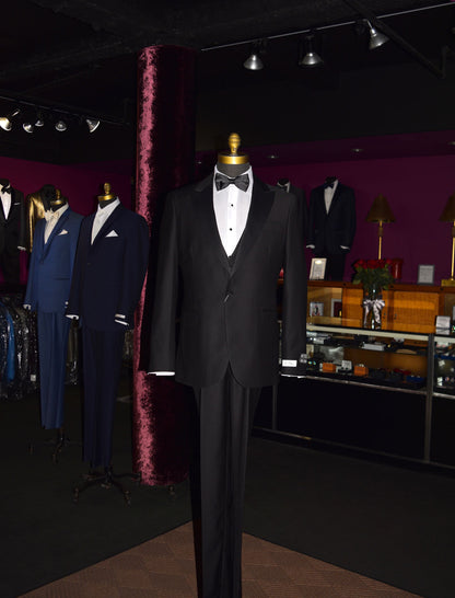 Black slim-fit 3 piece tuxedo on a mannequin in a formal wear store with other suits in the background.