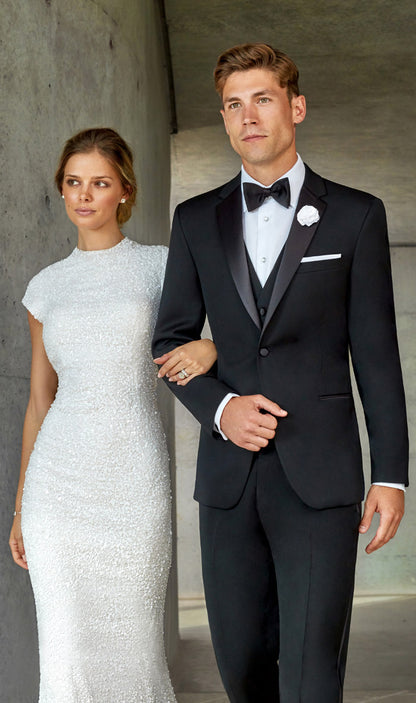 Man in a black notch lapel tuxedo and woman in a white dress standing against a neutral background