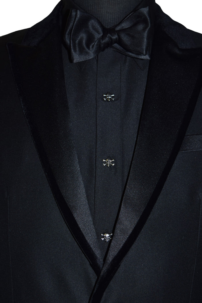close-up black tuxedo with velvet trim on lapel with a black shirt wearing skull and bones buttons and a black bowtie against a white background