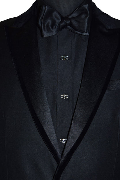 close-up black tuxedo with velvet trim on lapel with a black shirt wearing skull and bones buttons and a black bowtie against a white background