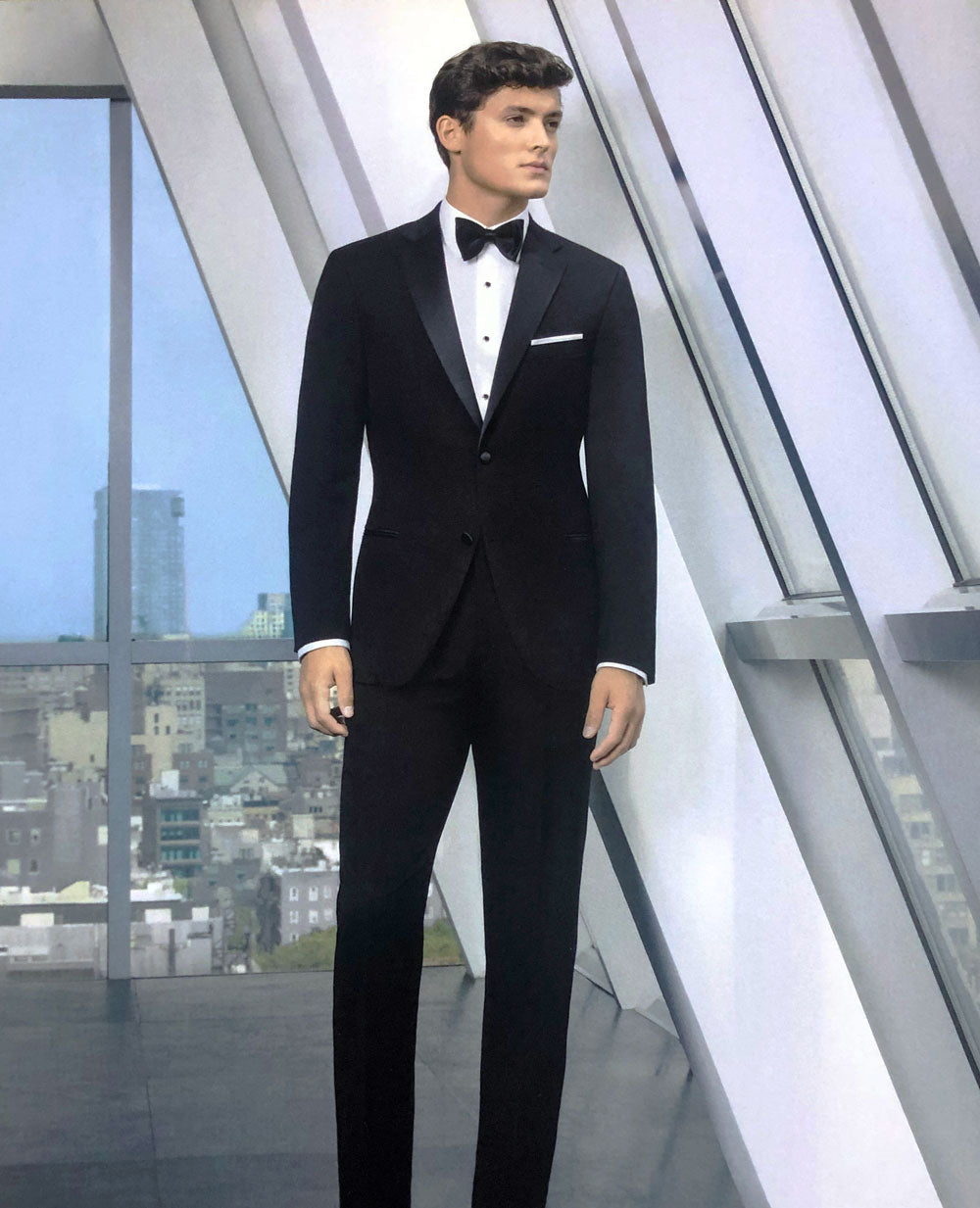 Model wearing a black fitted, 2 button notch lapel tuxedo with black bowtie in a modern building