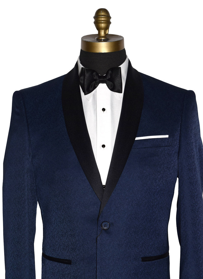 close-up front view-Navy blue tuxedo with black shawl collar with a brocade print on a mannequin with black tuxedo pants and a white tuxedo shirt and black satin bowtie in front of a white background