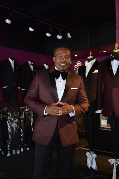 Man in a burgundy tuxedo standing in a tuxedo shop with tuxedos on mannequins.