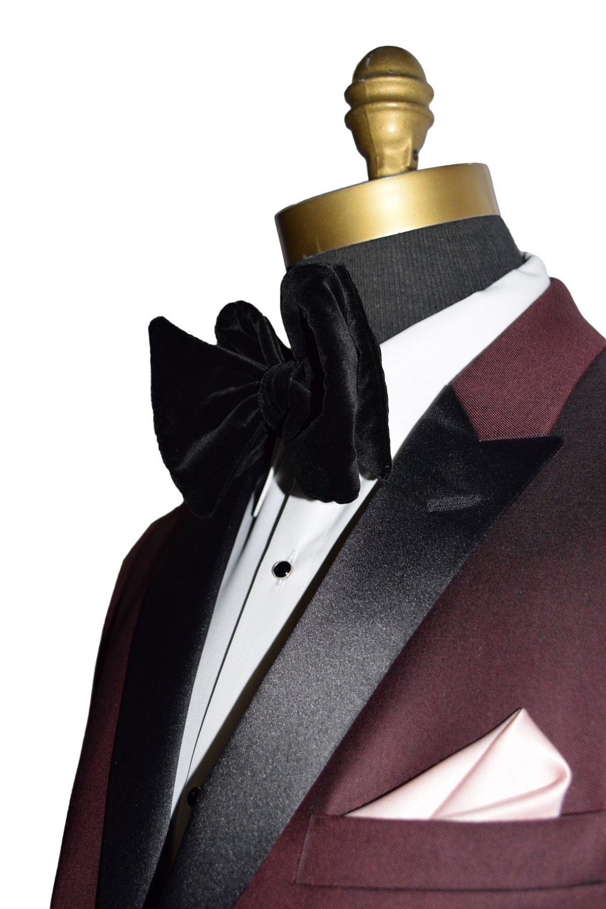 close up side view of burgundy tuxedo with black peak lapels shown with white tuxedo shirt and large black velvet bowtie on a mannequin against a white background